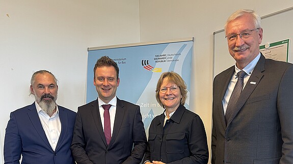 Drei Männer in Anzügen und eine Frau mit Bluse stehen vor einem hellblauen Aufsteller und blicken in die Kamera.