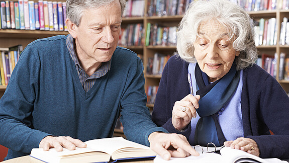Eine alte Frau und ein alter Mann sitzen an einem Tisch und lesen zusammen ein Buch.