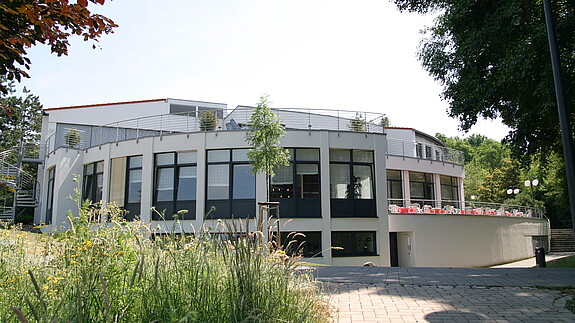 Pflegeseminar der dbb bundesseniorenvertretung Ein großes, helles, rundliches Haus ist im Mittelpunkt des Bildes. Im linken Vordergrund ist eine hochwachsende Wiese zu sehen.