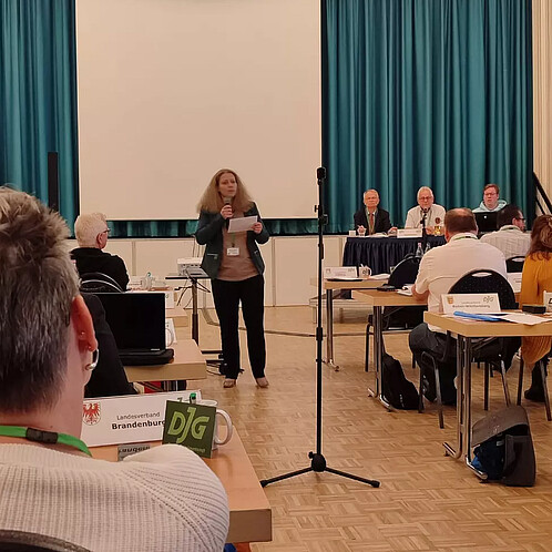 Eine Frau steht in der Mitte eines Seminarraums und spricht vor den anderen Anwesenden in ein Mikrofon
