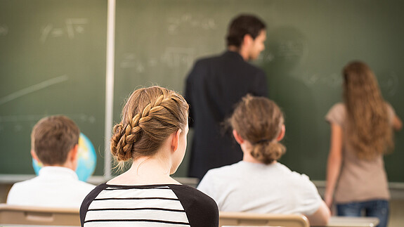 Das Foto zeigt einen Lehrer, der etwas an die Tafel schreibt, im Vordergrund sind die Hinterköpfe von Schülerinnen und Schülern zu sehen.