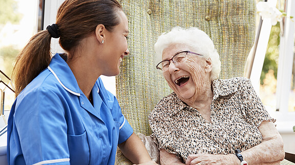 1. Oktober: Internationaler Tag der älteren Menschen Szene aus Seniorenresidenz: Eine Seniorin sitzt auf einer Bank, neben ihr eine Pflegefachkraft. Beide lachen.
