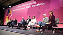 Menschen sitzen auf Stühlen auf einer Bühne und diskutieren miteinander. 
