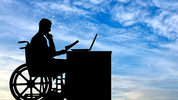 Die Silhouette eines Mannes im Rollstuhl, der an einem Schreibtisch sitzt und arbeitet.